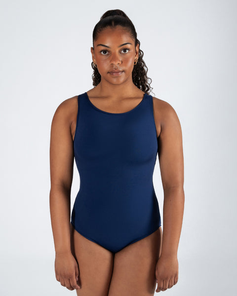 A woman modeling a sleeveless navy blue one-piece swimsuit against a light gray studio background.