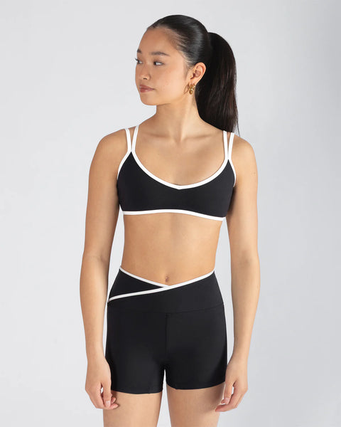 Woman wearing a black sports bra and high-waisted shorts with white straps on a light gray background